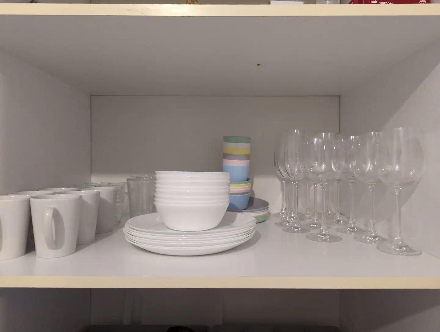 Une étagère avec des assiettes blanches, des tasses empilées et des verres à vin transparents.