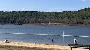 Vista de un lago con playa arenosa, una banca y gente cerca del agua.