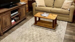 Un salón acogedor con detalles de madera, un televisor de pantalla plana sobre un soporte de madera, un sofá color beige, una mesa de centro de madera y una mesita auxiliar con una lámpara.