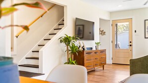 A modern living room with a staircase, wooden flooring, a flat-screen TV mounted on the wall, a wooden cabinet, and a door leading outside. - Hickory House - Family/Group and Pet-Friendly Retreat Near Lockhart Square! (Lockhart)