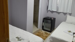 A small, compact hotel room with a bed, a small refrigerator, a bathroom with a shower, and a window with curtains. - ITIS HOTEL (Dom Eliseu)