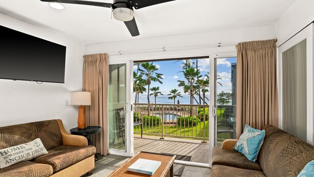 A modern living room with a sofa, a coffee table, and a TV. It features a sliding glass door leading to a balcony with a view of palm trees and a pool. - 3 Bedroom Ocean Front Condo with Pool & Elevator! (Kapaʻa)