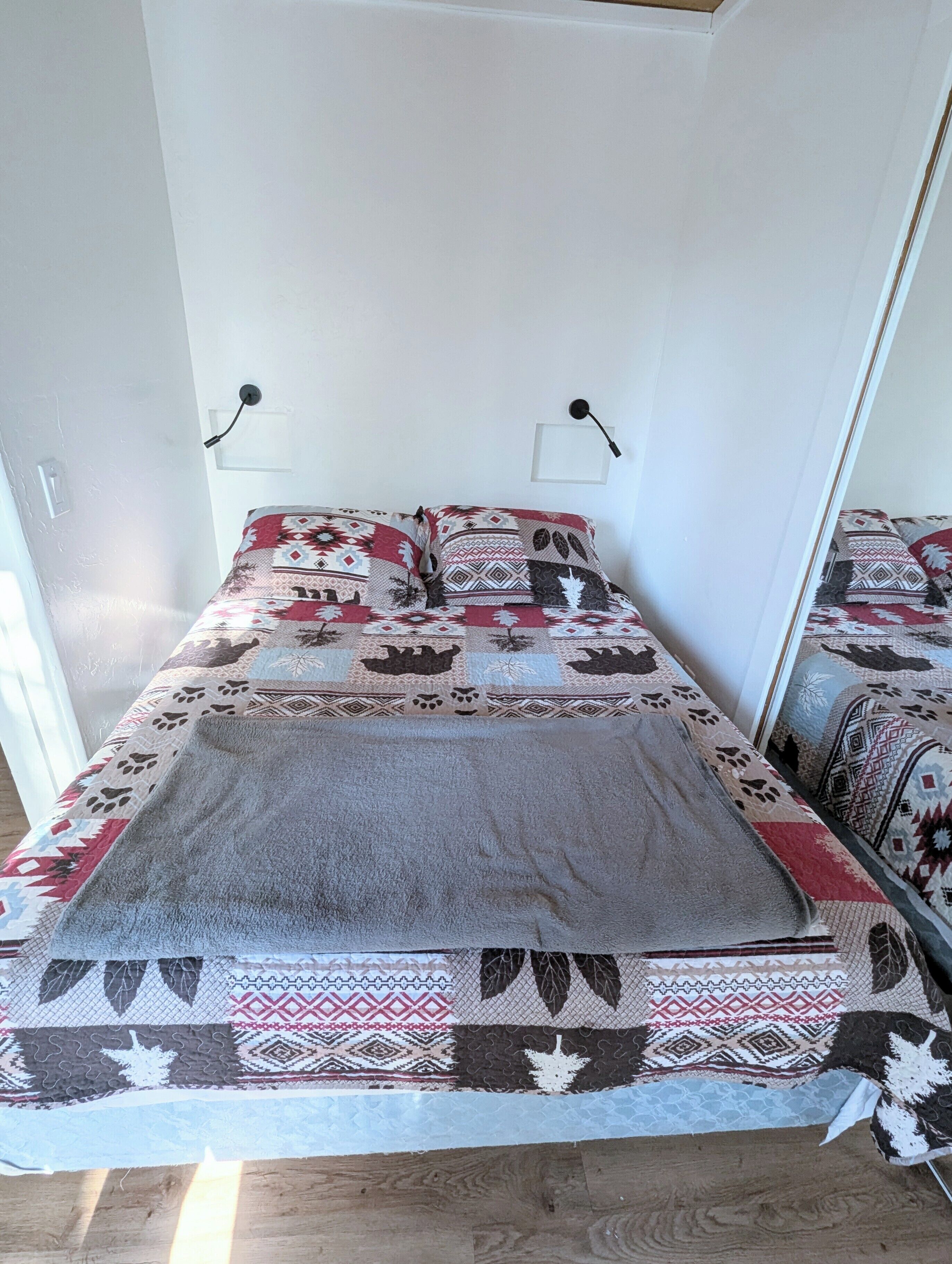 A bed with patterned bedding and a grey blanket, a white wall, and two wall-mounted lamps.