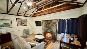 A cozy living room with a stone fireplace, wooden beams, a sofa, a coffee table, and a dining table with wine glasses.
