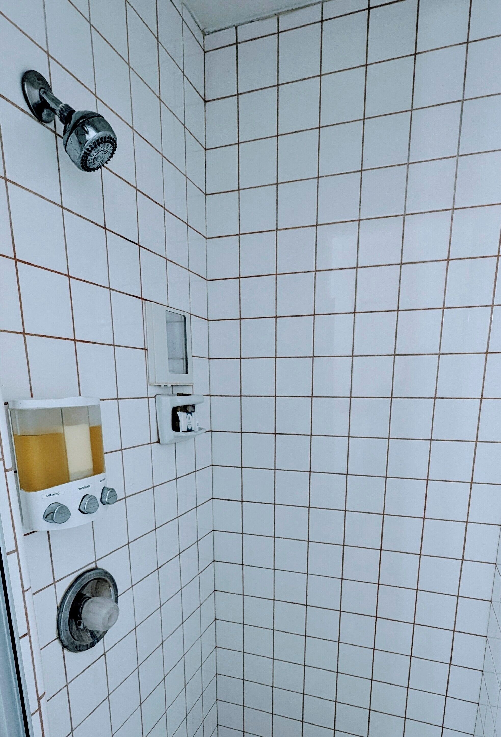 Un bagno con pareti in piastrelle bianche, un soffione della doccia, un erogatore di sapone e un piccolo specchio.