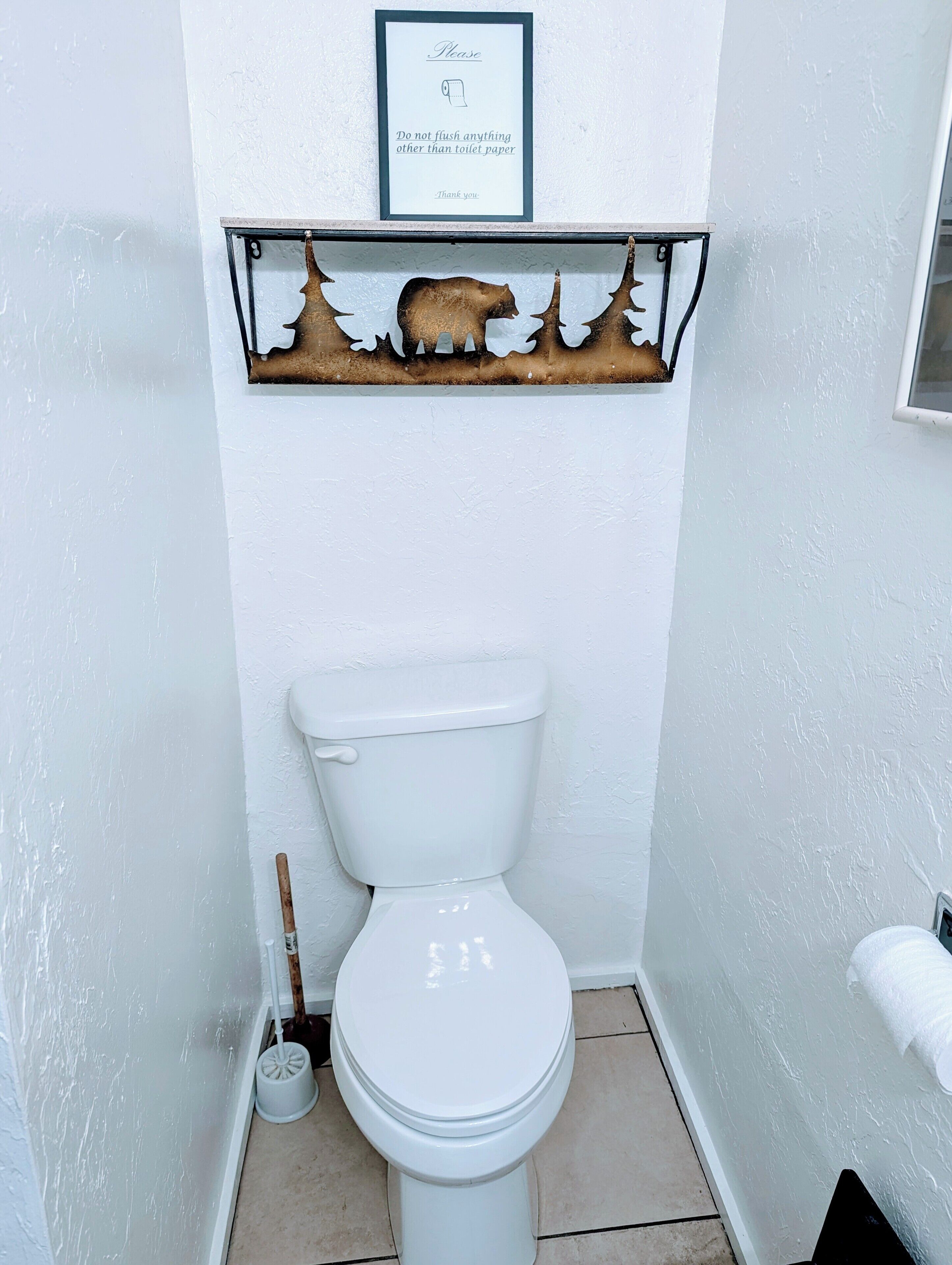 Un baño con inodoro blanco, un cartel enmarcado sobre el inodoro y un estante decorativo con un diseño de oso.