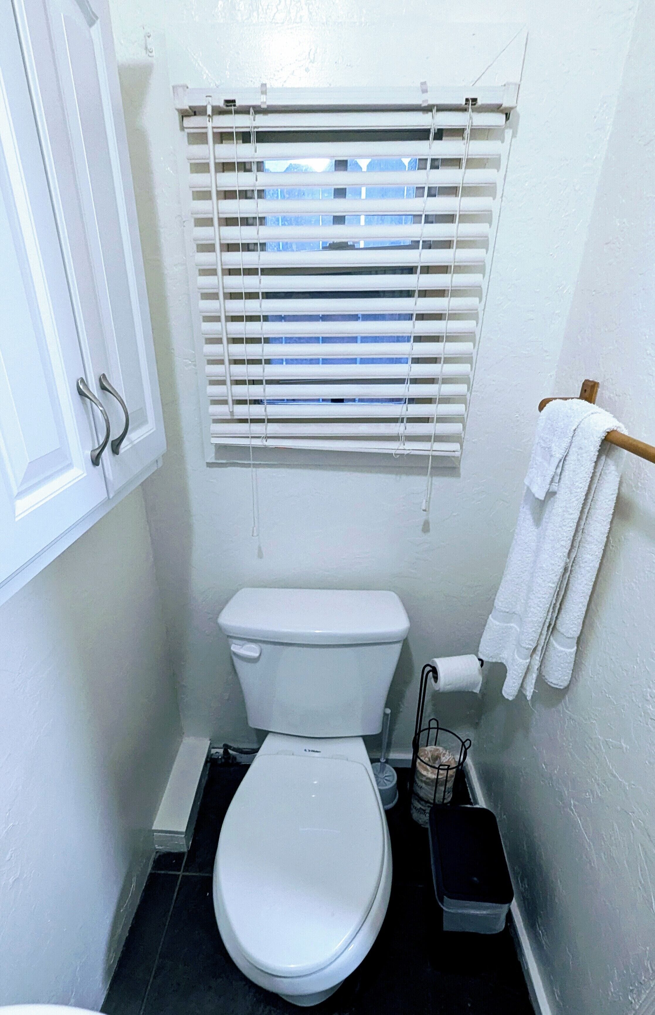 A small, white bathroom with a toilet, a window with blinds, a towel rack, and a trash bin.