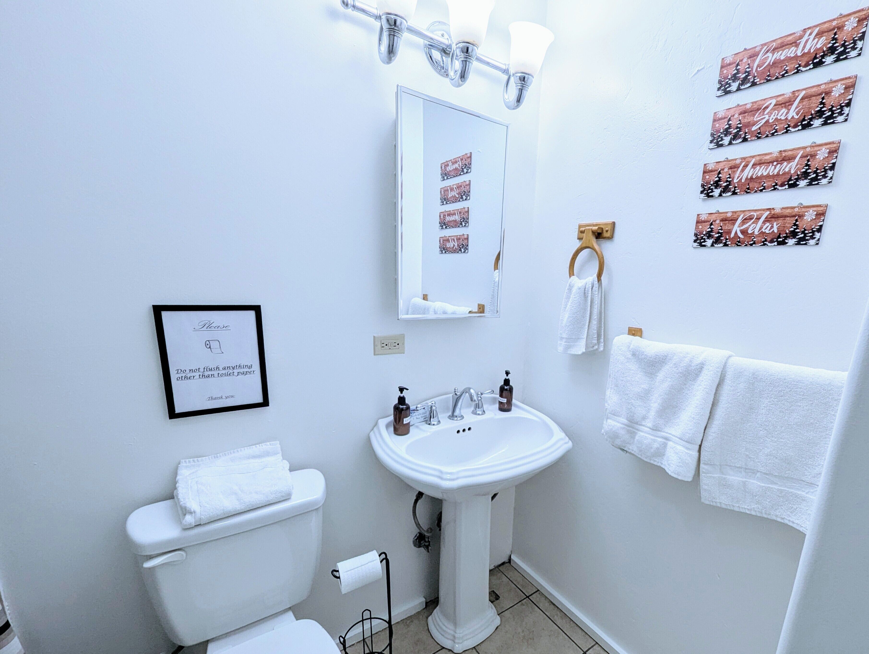 A bathroom with a white sink, a toilet, and a mirror. There are towels hanging on a rack and a framed sign on the wall.