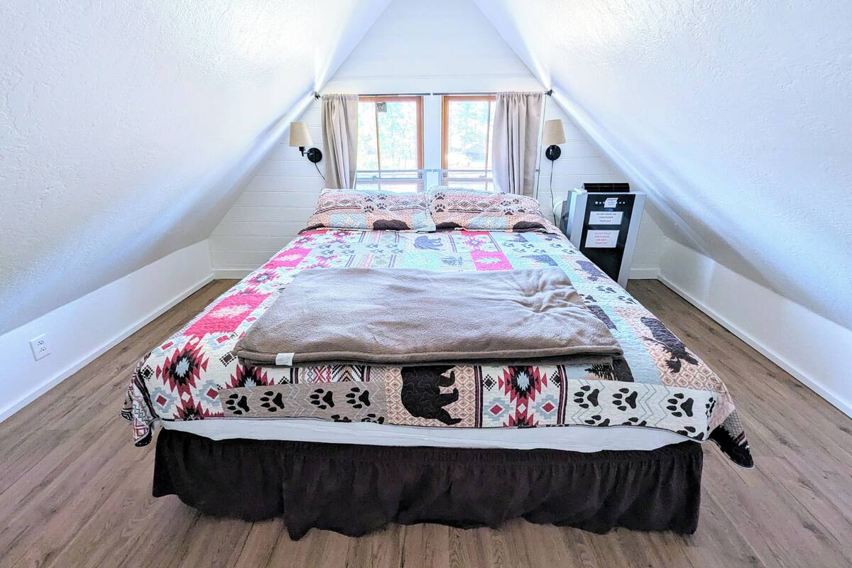 Une chambre avec un plafond en pente, un lit recouvert d’une couvre-lit à motifs, une fenêtre avec des rideaux et un radiateur fixé au mur.
