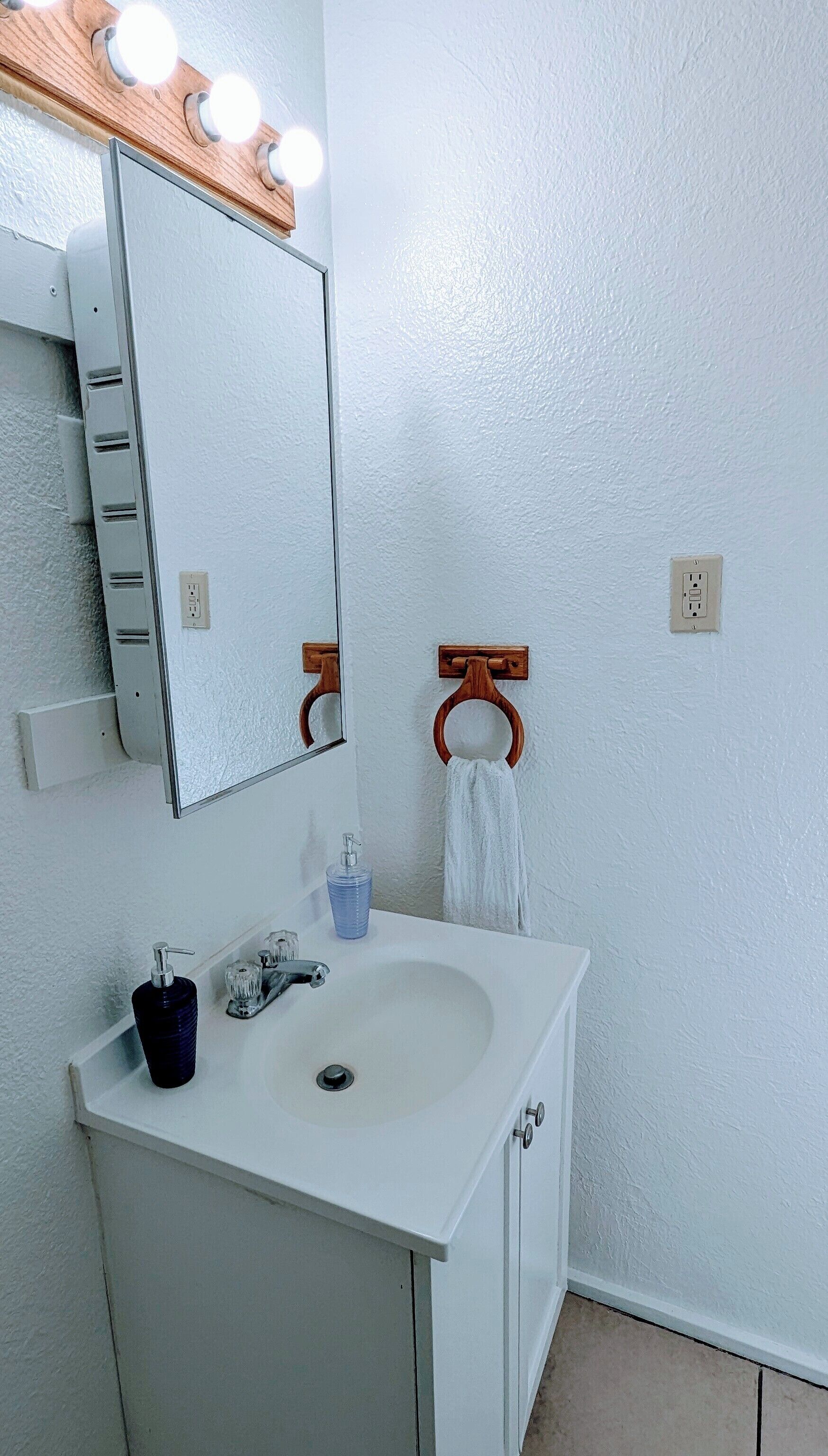 A bathroom with a white sink, a mirror with lights, a towel rack, and a soap dispenser.