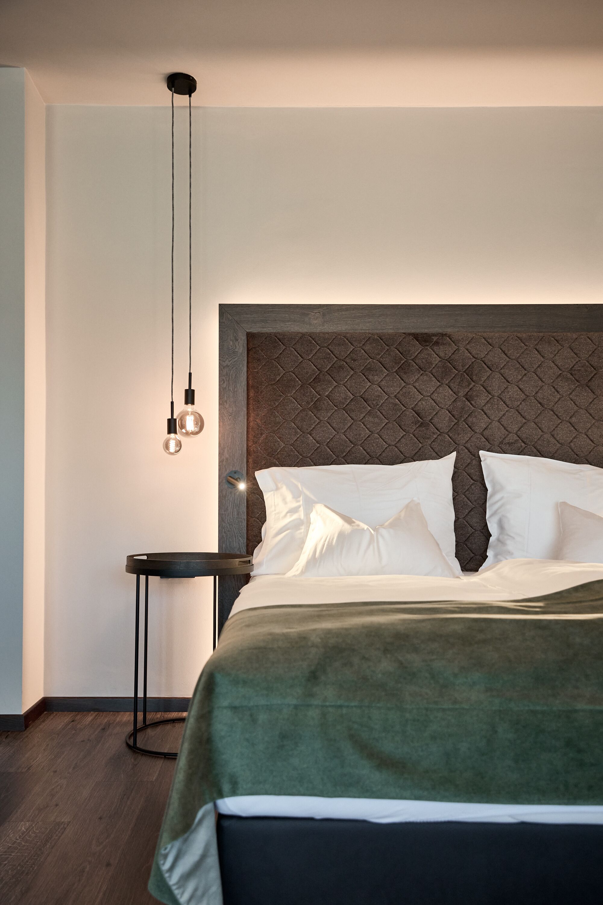 A neatly made bed with white linens and a green throw blanket, a round side table with a black base, and a pendant light fixture above the bed.