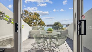 A balcony with a glass door, a round table with chairs, a potted plant, and a view of a lake and trees. - Horizons 404 (Jindabyne)