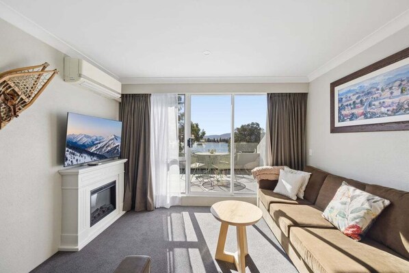 A living room with a fireplace, a television, a sofa, a small table, and a balcony with a view of a lake and mountains. - Horizons 404 (Jindabyne)
