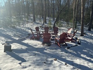Ein verschneiter Außenbereich mit mehreren roten Adirondack-Stühlen, die kreisförmig angeordnet sind, einer Feuerstelle und einem Steinkamin.
