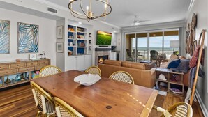 A spacious living area with a wooden dining table, wicker chairs, a fireplace, and a view of the ocean through large windows.