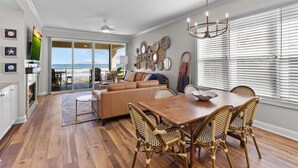 A spacious living area with a wooden floor, a large sofa, a dining table with wicker chairs, and a view through large windows.