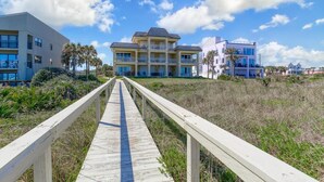 A wooden boardwalk leading to a multi-story building with balconies, surrounded by palm trees and a clear sky.