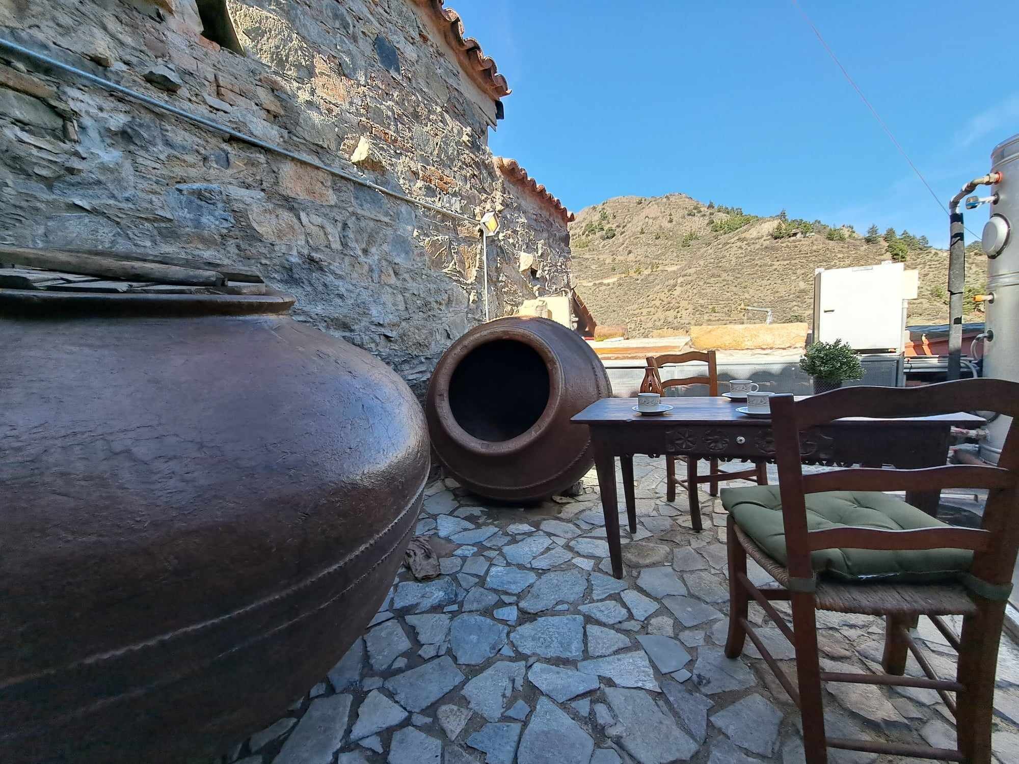 A stone patio with large clay pots, a wooden table with chairs, and a view of a hillside.