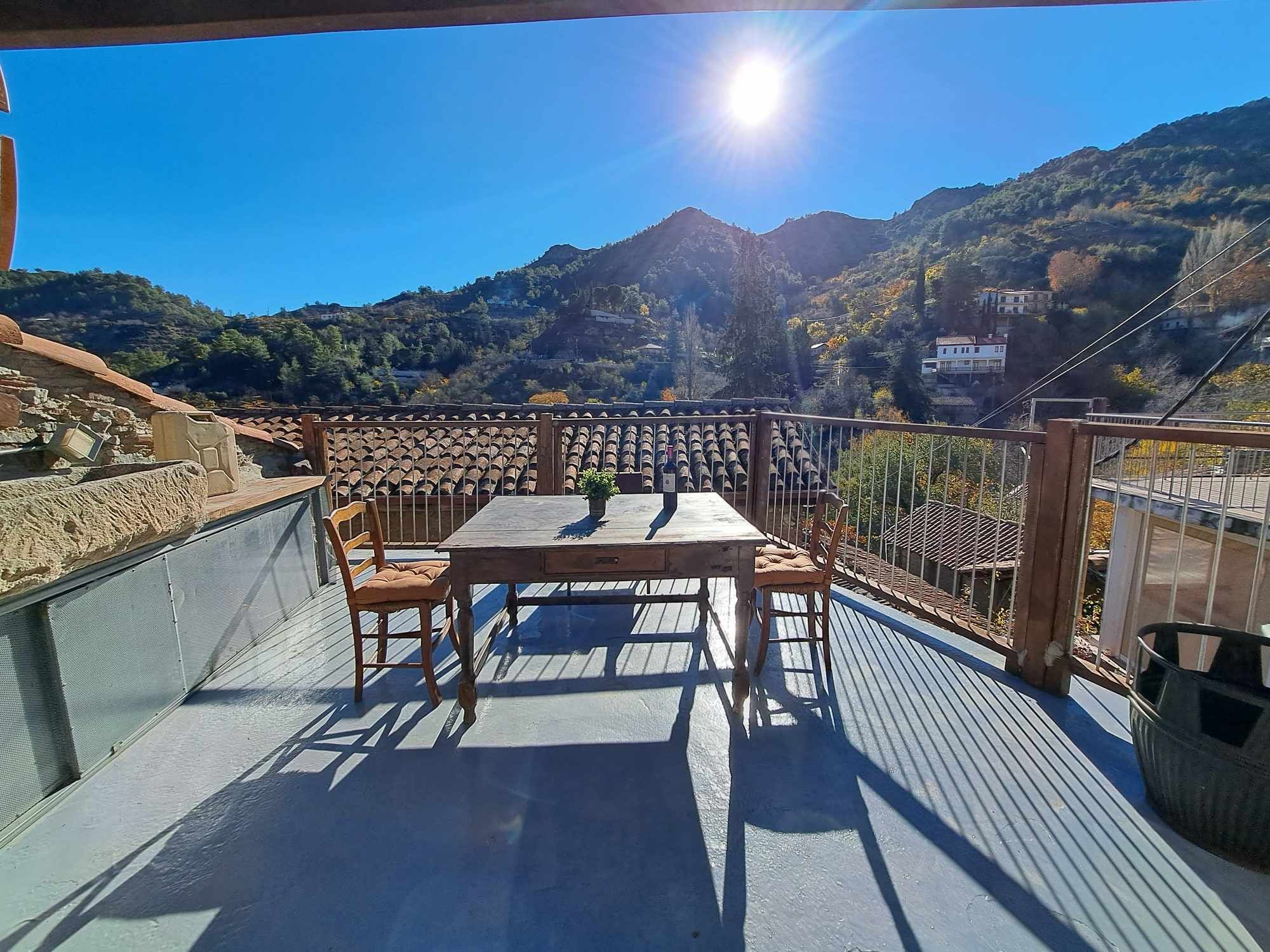 A terrace with a table and chairs, overlooking a valley with mountains and houses.
