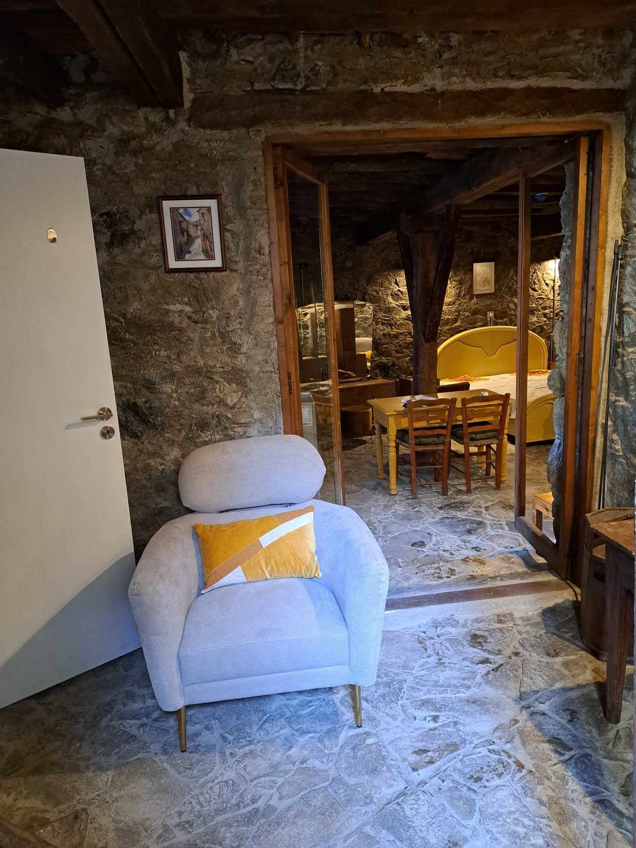 A rustic room with a stone wall, a white armchair with a yellow cushion, a wooden table, and a yellow bed.