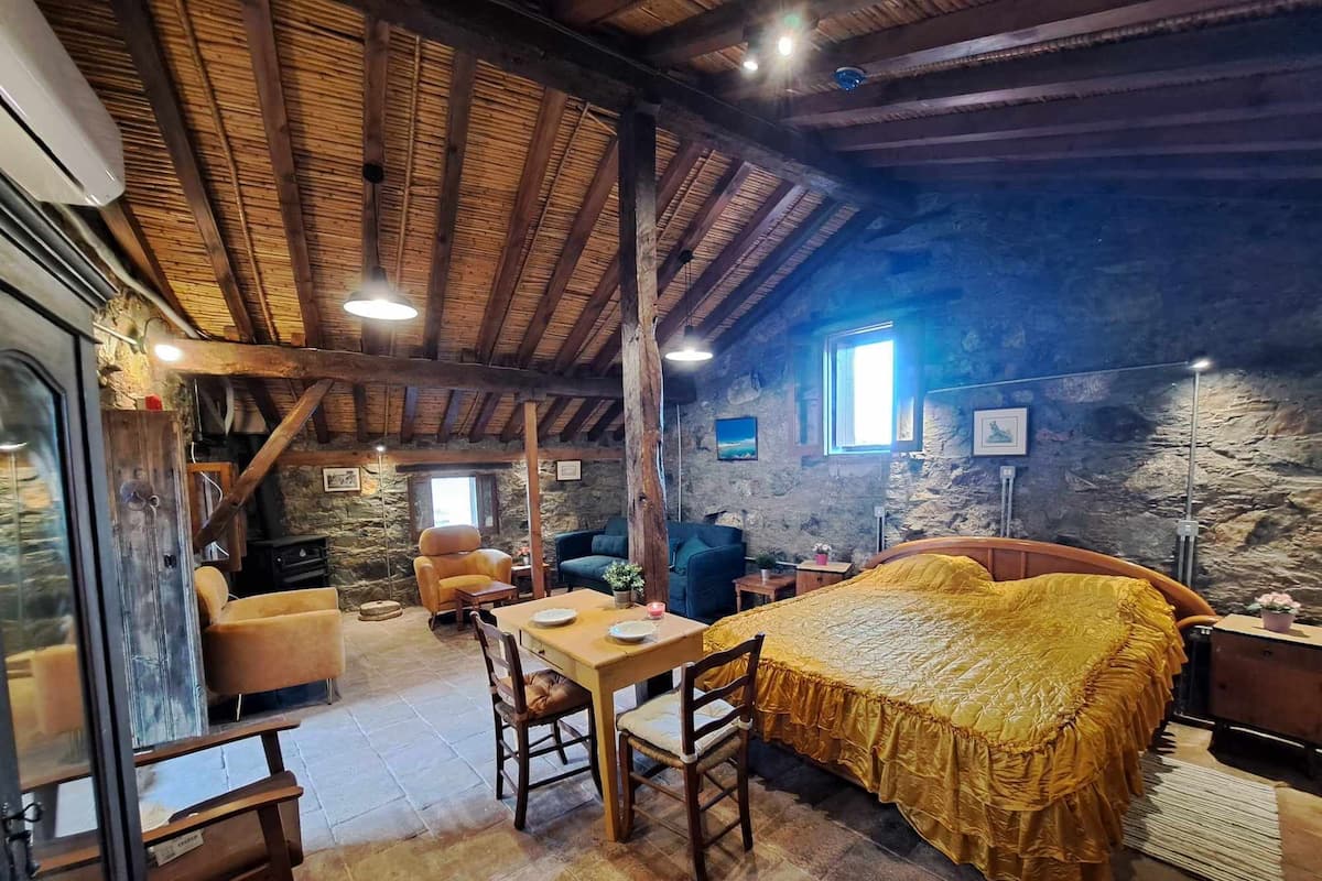 A rustic room with a bed, a table, and chairs. There is a stone wall and wooden beams on the ceiling.