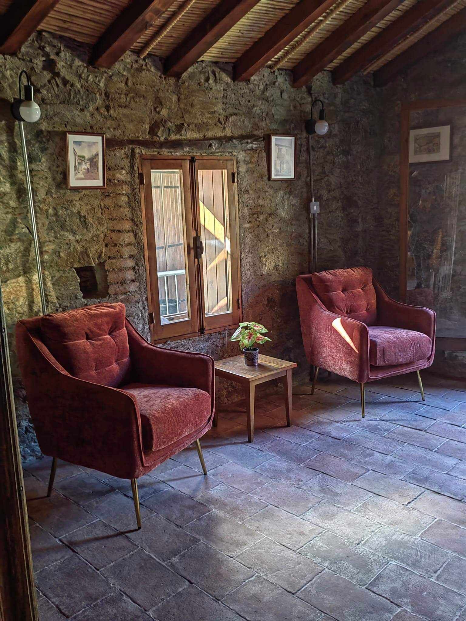 A rustic room with stone walls, wooden beams, two red velvet armchairs, a small wooden table with a potted plant, and framed pictures on the walls.
