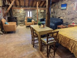 Una habitación rústica con paredes de piedra, vigas de madera, chimenea de piedra, un sofá, una silla, una mesa y una cama con colcha amarilla.