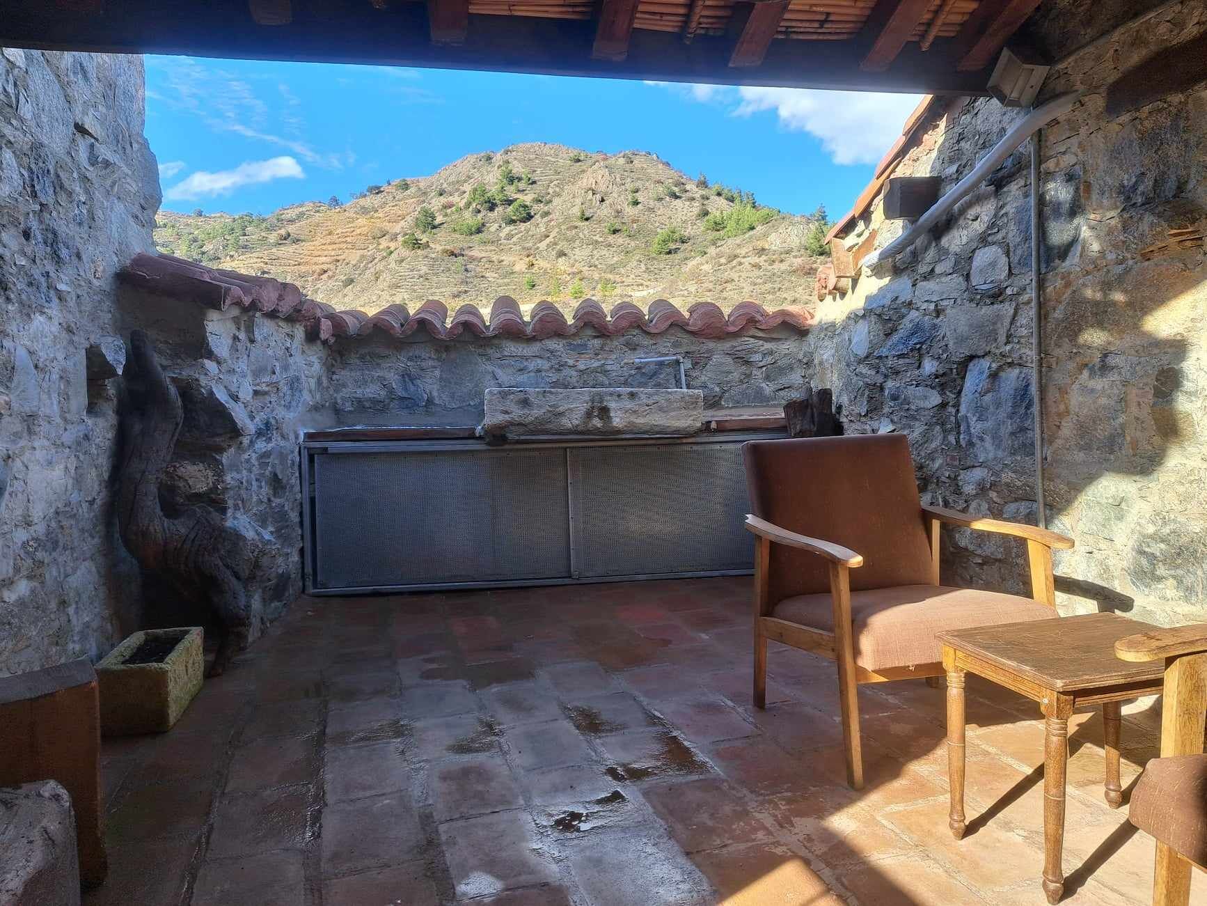 Un patio al aire libre con paredes de piedra, una mesa y una silla de madera, un banco de piedra y vistas a una colina.