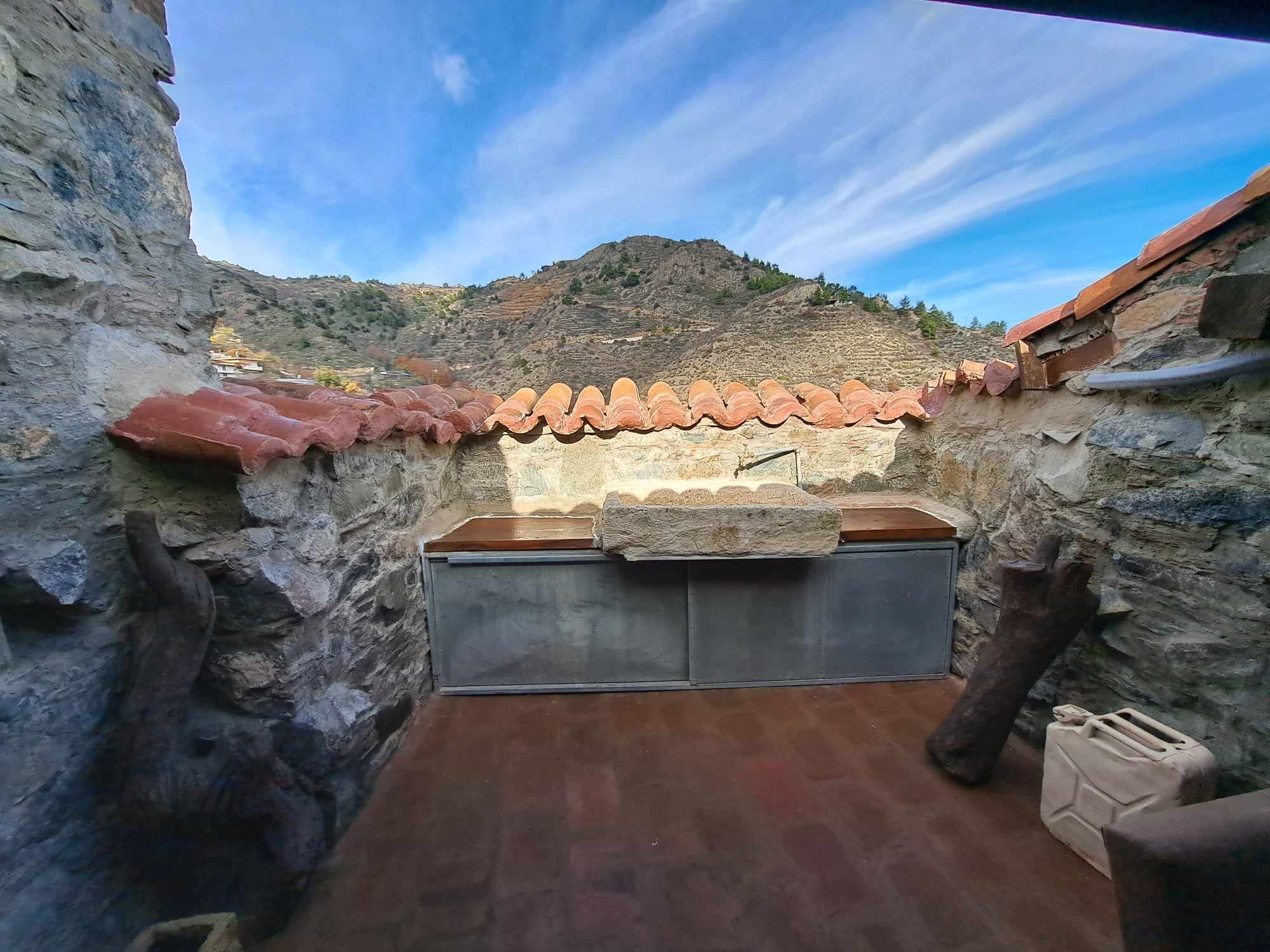 A stone-walled outdoor kitchen with a tiled roof, a stone bench, and a view of a mountainous landscape.