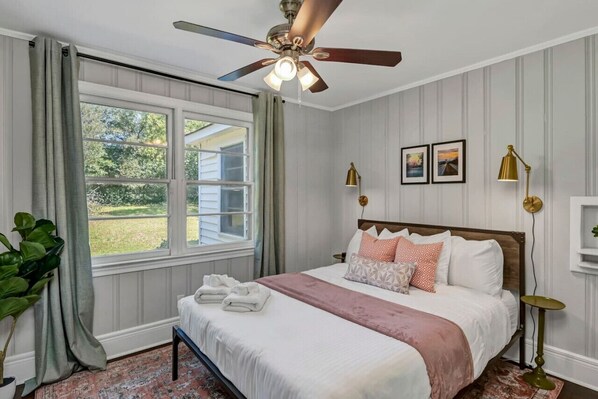 A bedroom with a bed, a ceiling fan, a window with curtains, a bedside table, and a lamp.