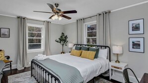 A bedroom with a bed, a nightstand, a chair, a desk, a ceiling fan, and two windows with curtains. - The Duke | 3BR 1 BA | Pet Friendly! (Columbia)