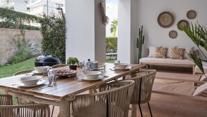 An outdoor dining area with a wooden table set for four, surrounded by wicker chairs. A barbecue grill is visible in the background, and a sofa with cushions is set up against the wall. - Casa Livia with Auping beds (Estepona)
