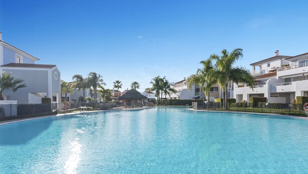 A clear blue swimming pool surrounded by white buildings with palm trees, a covered area, and a lifeguard stand. - Casa Livia with Auping beds (Estepona)