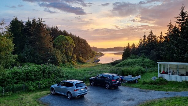 A gravel path leading to a cabin with a trailer, surrounded by dense forest and a lake at sunset. - Vollvaagen Apartments Smøla (Smøla)