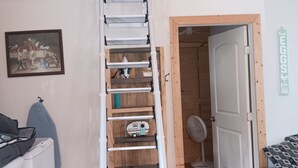 Une chambre en mezzanine accessible par une échelle en bois, avec un espace de couchage surélevé, une petite table avec une chaise et une porte donnant sur une autre pièce.