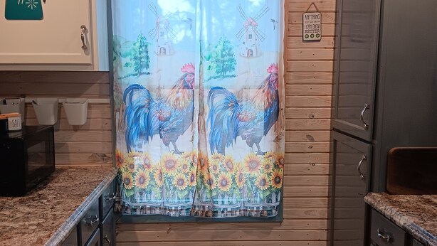 Une cuisine avec des meubles en bois, une fenêtre ornée d’un rideau représentant un coq et des tournesols, et une pancarte sur laquelle on peut lire : « Restez calme et mangez des carottes ».