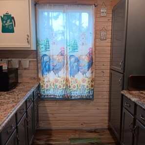A kitchen with wooden cabinets, a window with a rooster and sunflower curtain, and a sign saying "Romaine Calm and Carrot on."