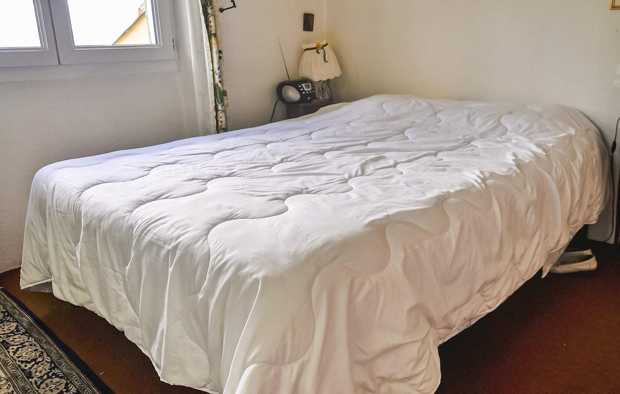A single bed with a white duvet in a room with a window, a lamp, and a small table with a telephone.
