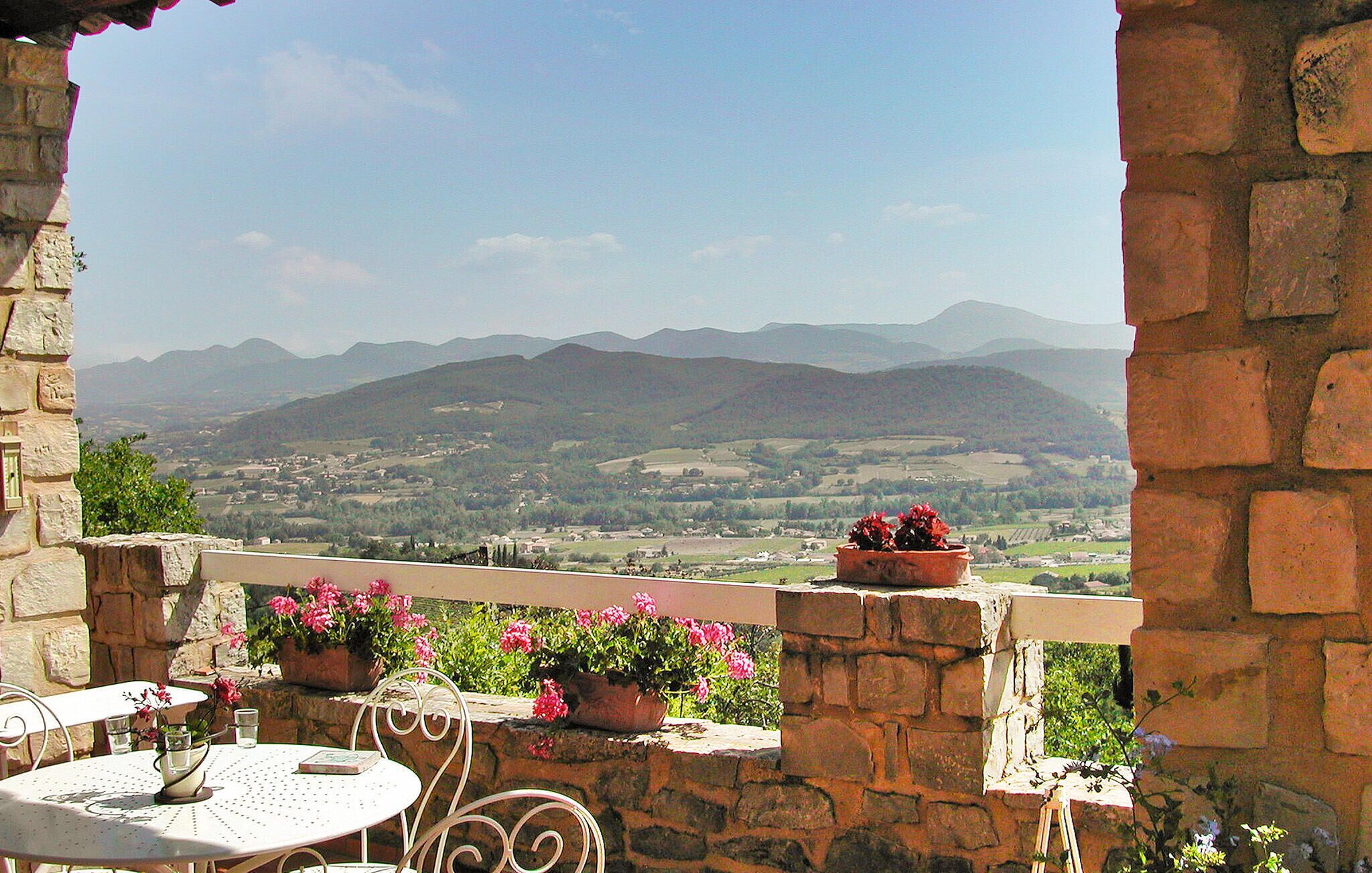 A stone balcony with a table and chairs, potted plants, and a view of distant mountains.