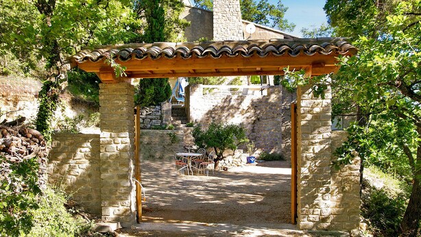 Una pérgola de piedra con techo de tejas, que conduce a un muro de piedra y un edificio de piedra con chimenea al fondo.