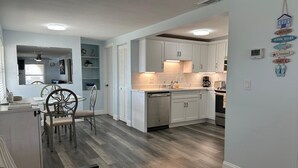 A modern kitchen with white cabinets, a dining area with a glass table and chairs, and a living room with a sofa and a coffee table. - Crescent Beach Retreat - Steps to Sand - Pet Friendly (St. Augustine)