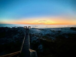 Ein hölzerner Steg führt zu Strandliegen an einem Sandstrand, im Hintergrund ist ein Sonnenuntergang zu sehen.