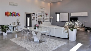 A modern living room with a white sofa, clear chairs, a glass coffee table, and a white rug. There are potted plants, a bookshelf, and a flat-screen TV mounted on the wall. - La Villa De Notre Dame (Chateaurenard)