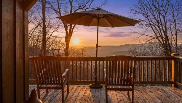 A wooden deck with two wooden chairs and a table, an umbrella, and a view of a sunset over a mountainous landscape. - Mountain Retreat, Sunset Views, and 20 Minutes to Asheville! (Swannanoa)