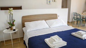 Et hotelværelse med en seng, et lille bord med en plante og vinglas, samt et spiseområde med stole.