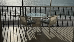 Un balcon avec une table en verre et deux fauteuils en osier, donnant sur une plage où l’on peut admirer les vagues et un ciel dégagé.