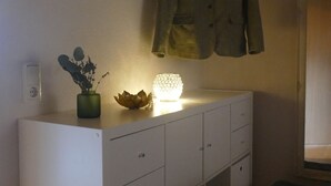 Un comò bianco con un vaso e una candela, un cappotto appeso sopra e una panca alla destra.