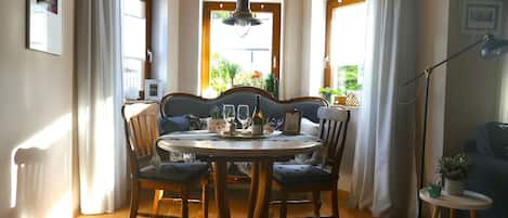A dining area with a round table, four chairs, and a chandelier. There are windows with curtains, a floor lamp, and a small table with a plant.