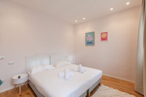 Een slaapkamer met een wit bed, twee kussens, een nachtkastje en twee kunstwerken aan de muur.