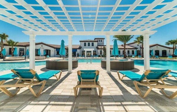A pool area with lounge chairs and umbrellas, a covered pergola, and a building in the background. - Lake view Windsor 4BD Disney (Kissimmee)